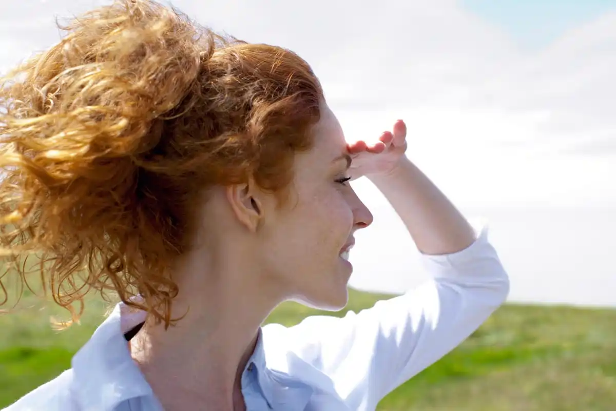 Eine Frau mit lockigem, rotem Haar steht in der Natur und schaut in die Ferne.